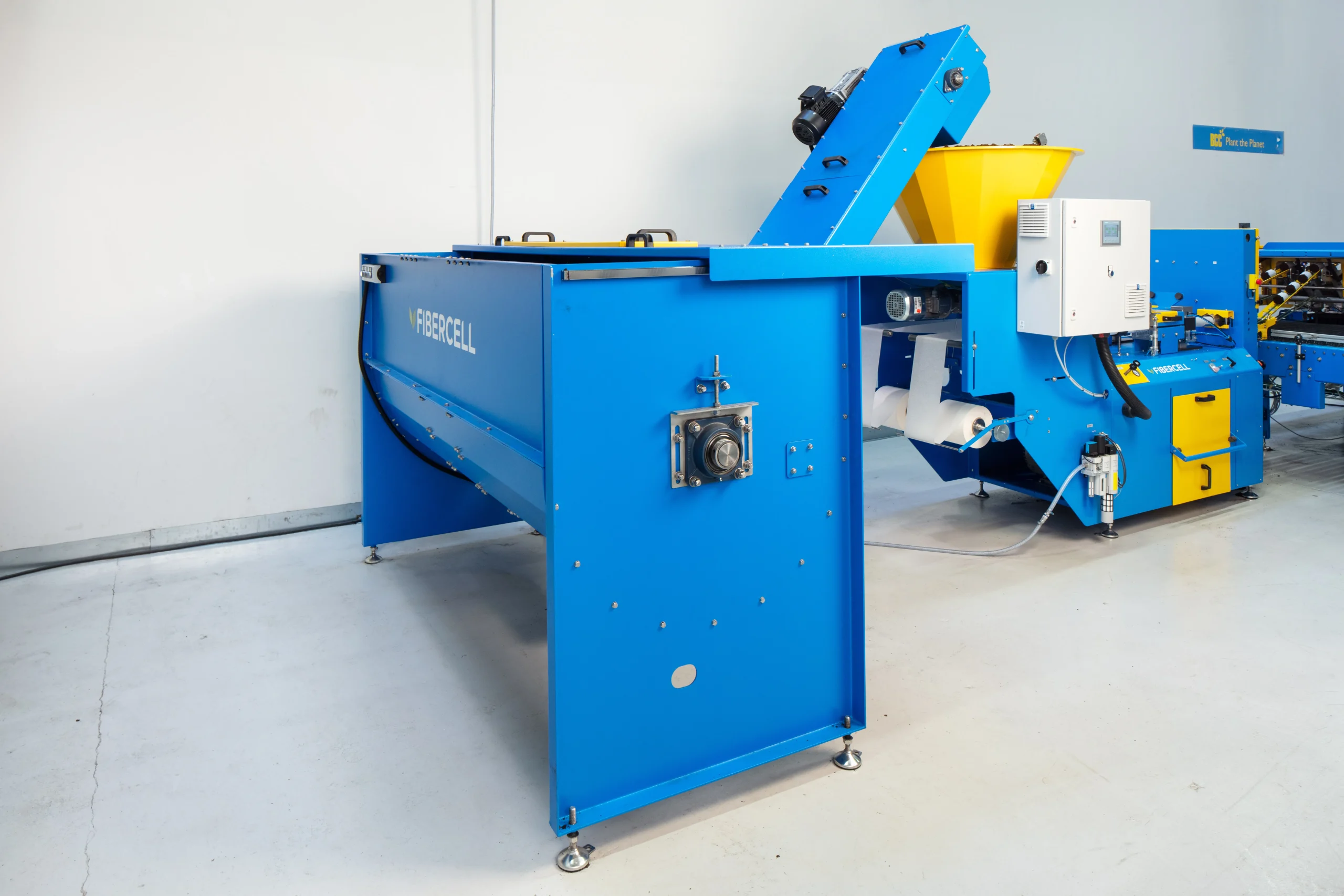 A blue industrial machine labeled "FIBERCELL" is positioned on a clean, light-colored floor. It has a conveyor system with a yellow funnel, surrounded by other machinery parts.