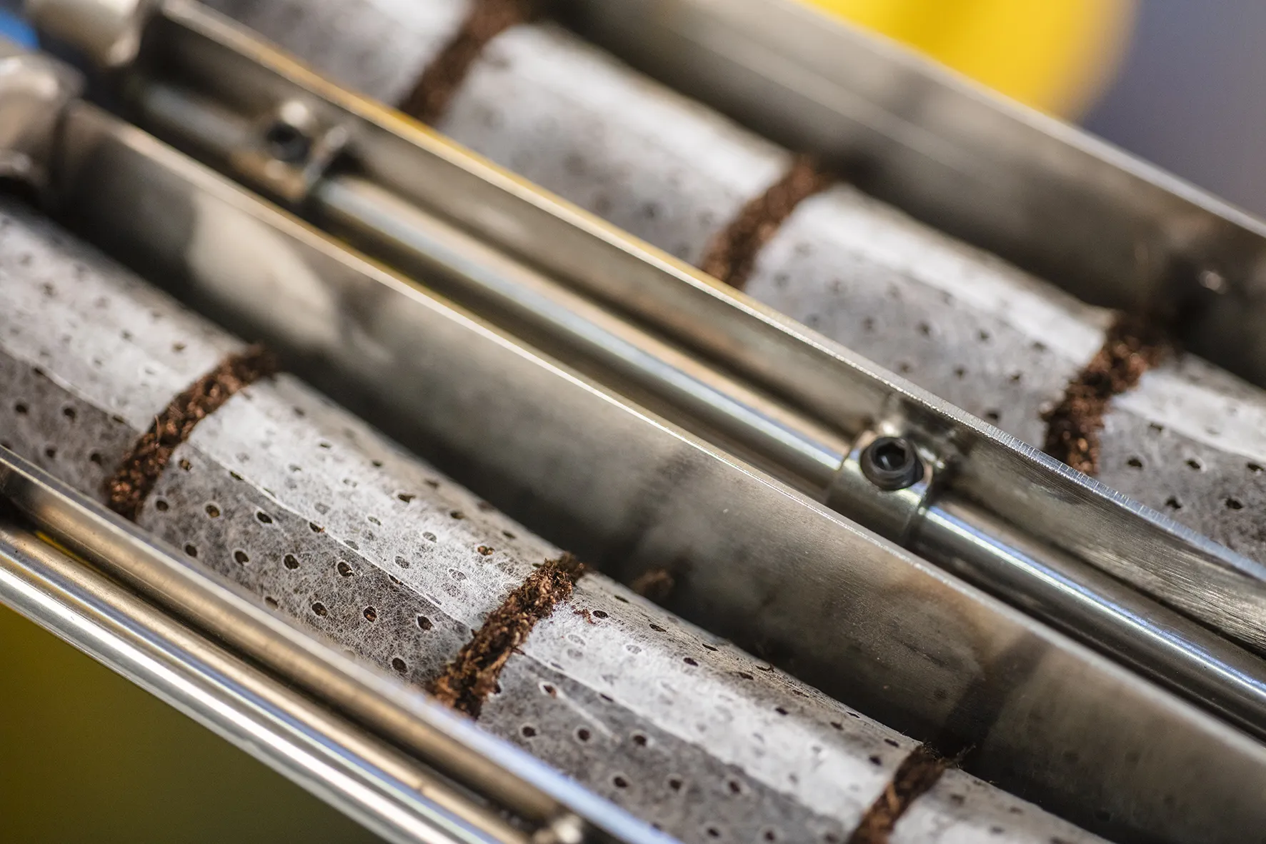 Metal cylinder perforating tea bags with visible tea leaves along seams, in a close-up industrial setting.