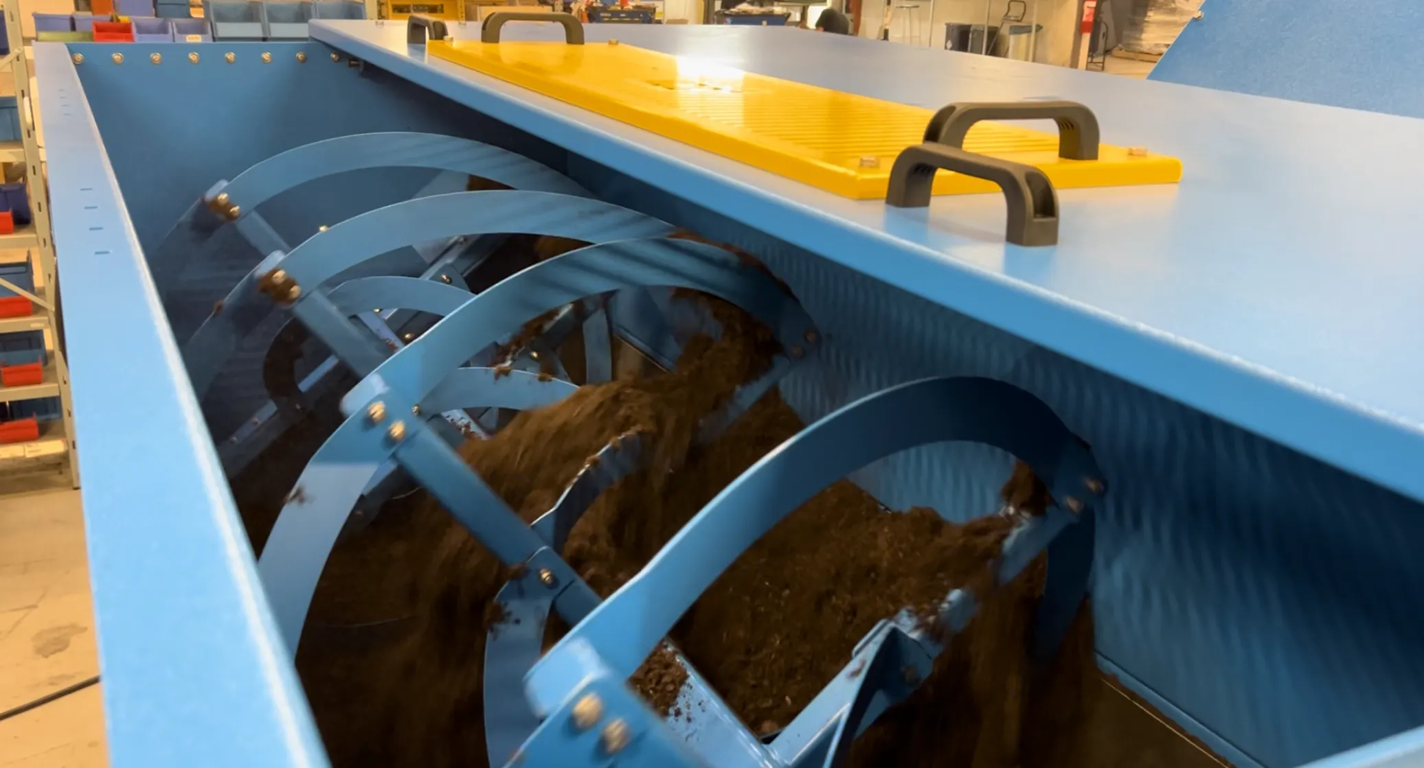 A large industrial mixer with blue, rotating blades churns dark, loose material inside a blue metal container. The setting is an indoor workshop with visible shelves and equipment.