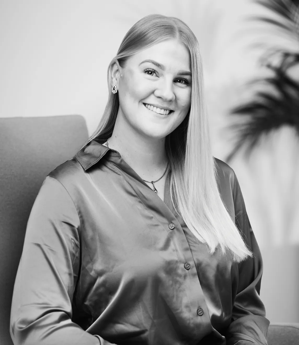 A person smiles while seated on a chair, wearing a shiny buttoned blouse, with potted plants softly blurred in the background.