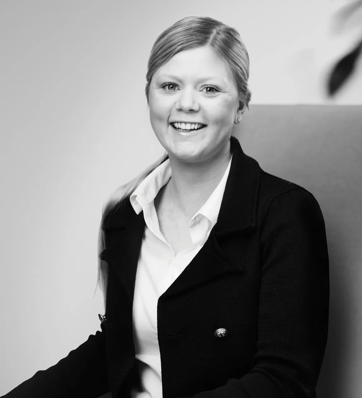 Smiling woman sits, wearing a white shirt and dark blazer, against a plain background. A blurred leaf is visible in the top right corner.