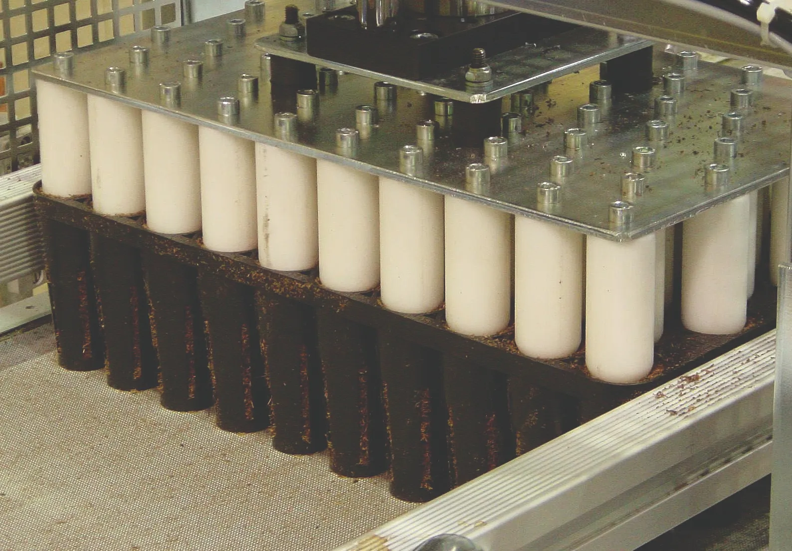 A mechanical device presses cylindrical white components into black holders within a metal framework, possibly part of an industrial or manufacturing process. The surrounding area features metal and grid structures.