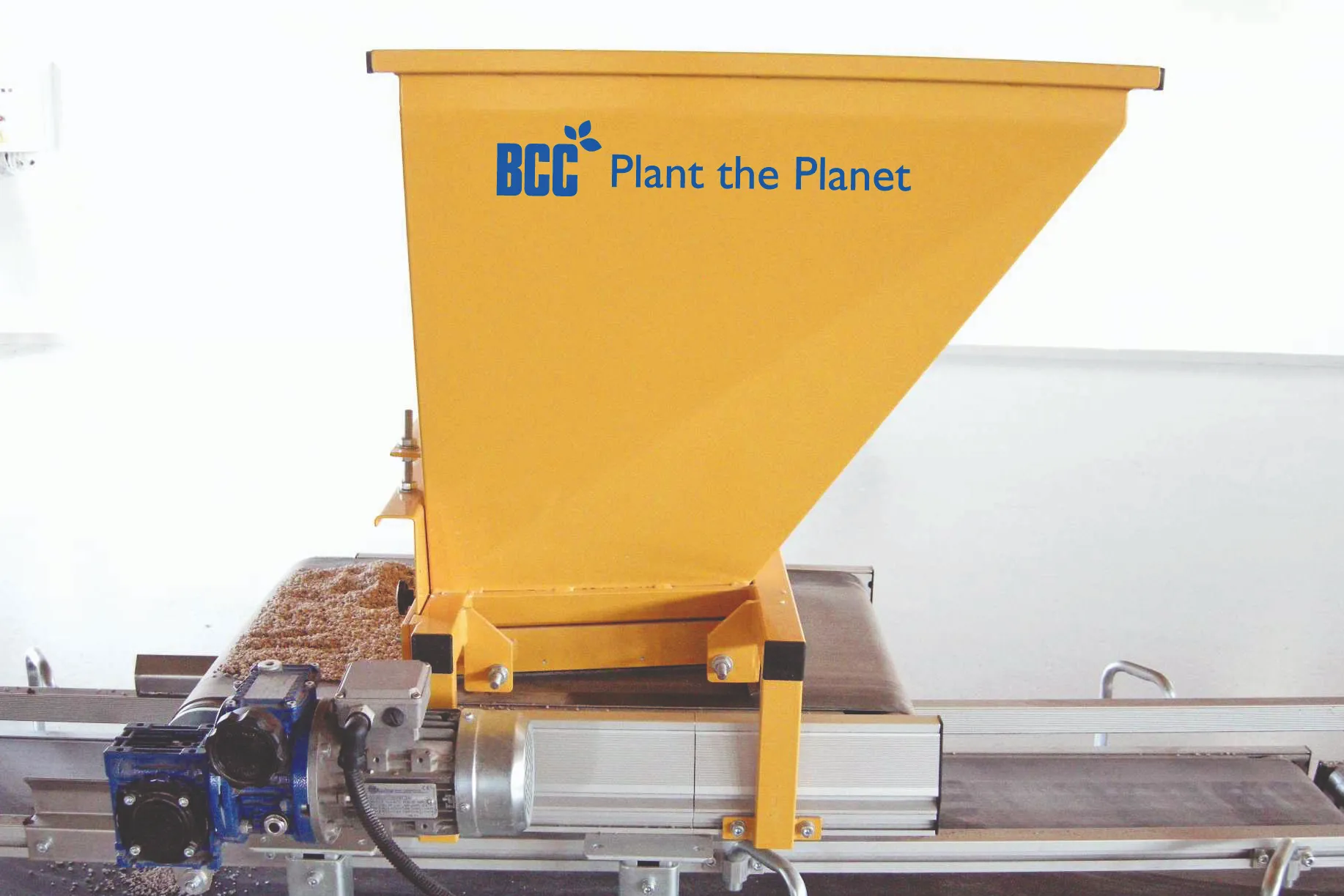 A yellow hopper dispenses seeds onto a conveyor belt, assisted by machinery in a clean, industrial setting. Text reads: "BCC Plant the Planet."