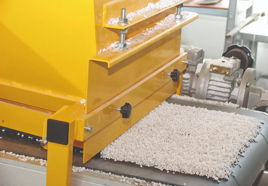 A yellow industrial machine dispenses granular material onto a moving conveyor belt in a processing facility, with mechanical components visible in the background.