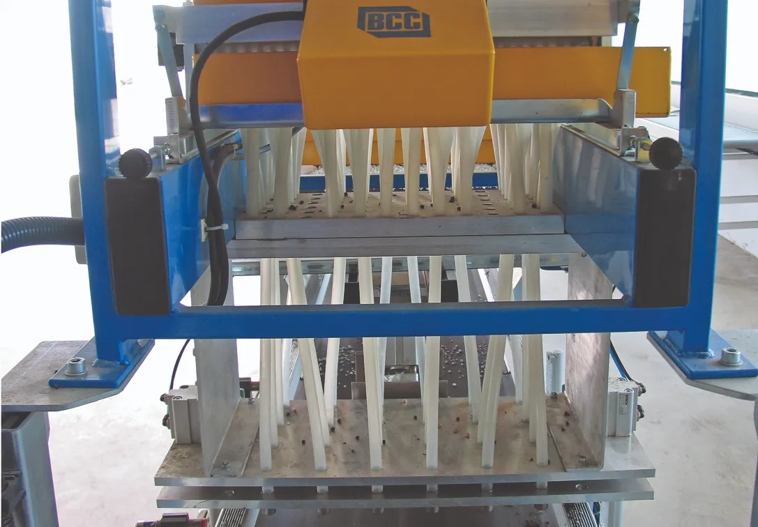 Machine with multiple vertical tubes pushing objects downward through a grid, surrounded by blue metal framework. A yellow component is marked with "BCC," in an industrial setting.